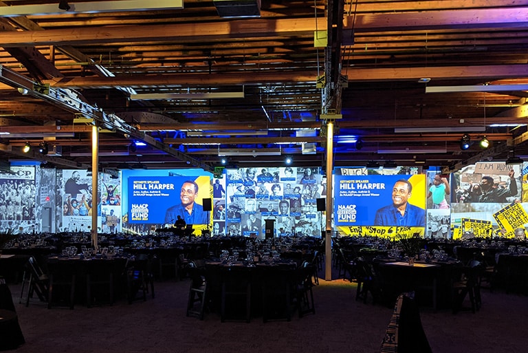 Tables are set and ready for the NAACP Freedom Fund Dinner. A collage of historic images creates the graphic across the large projection wall. Blue and yellow lighting throughout the space tie into the event colors.