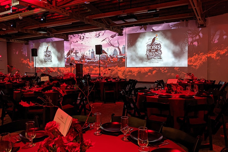Image showing the atmosphere of a nonprofit gala called Roses and Ravens. The projection wall displays a thematic image in dark reds and blacks, echoing the event's theme. Tables in the foreground are elegantly set with red linens and black place settings. The event logo is prominently projected on the wall in two places.