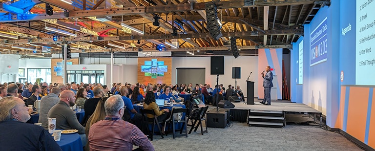 A presenter addresses the audience from the stage during a corporate meeting