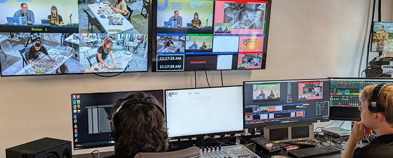 Two video technicians monitor a live hybrid event broadcast from a professional control room, with multiple screens displaying camera feeds and production software.
